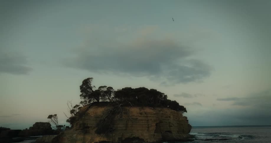 Waves crash onto a small coastal island plateau with trees on top in the early morning
