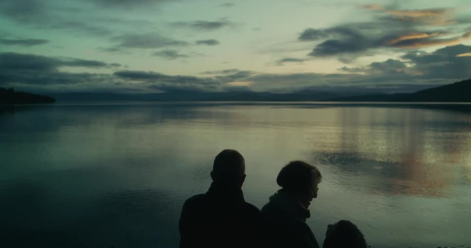 Silhouette of a couple and a dog in front of a lake that reflects the early morning sky