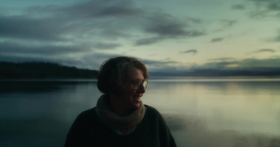 An individual stands in front of a lake smiling and talking