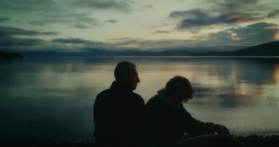 Two people sit by a tranquil lake at dusk silhouetted against the serene evening sky