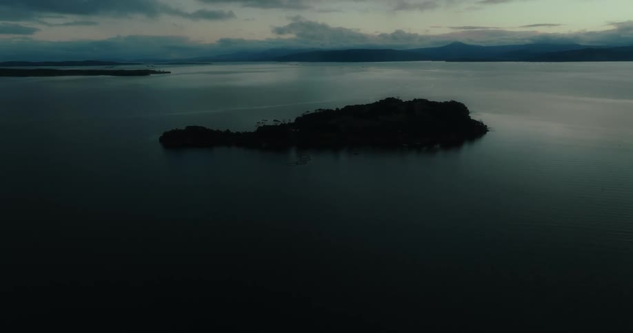 A small dark island sits in the middle of a calm reflective body of water at dusk