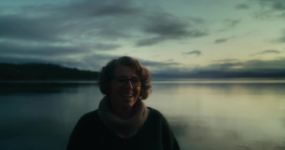 A woman wearing glasses and a thick scarf stands near a calm lake at dusk