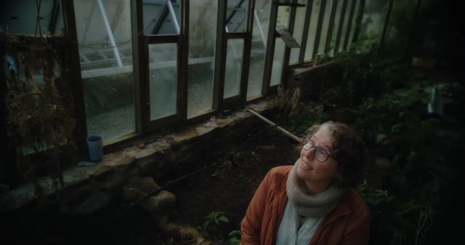 A woman wearing glasses and a thick scarf sitting inside a greenhouse filled with various plants