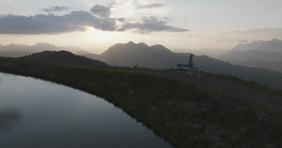 Woman with a dog on a leash walking next to a calm body of water with mountains in the background under a cloudy sky