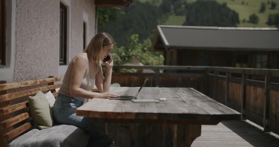 A woman sits on an outdoor bench talking on the phone and writing in a notebook with a scenic view of mountains and houses in the background