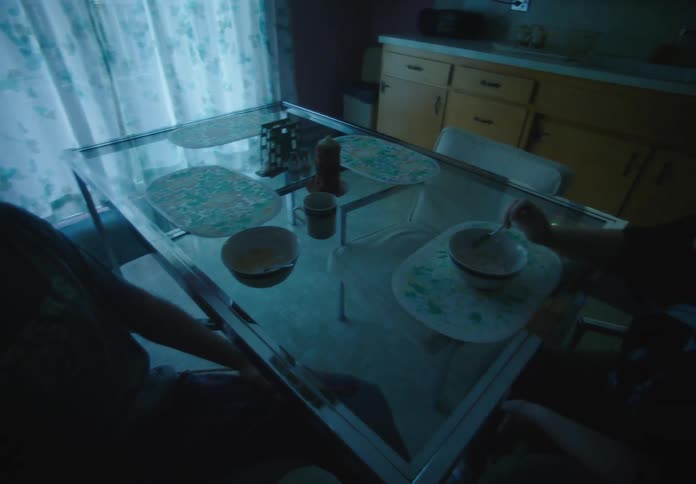 A man and a woman sit and talk at a kitchen table set for a meal illuminated by dim blue light as the woman smiles while listening