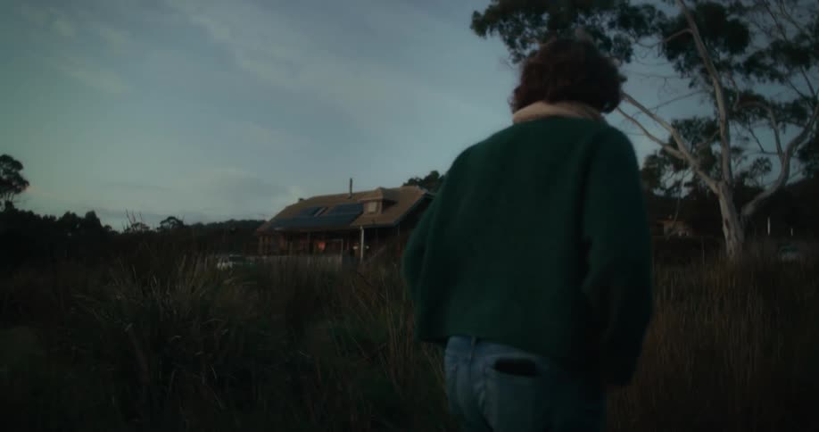 A man walking near a house surrounded by tall grass and trees during twilight