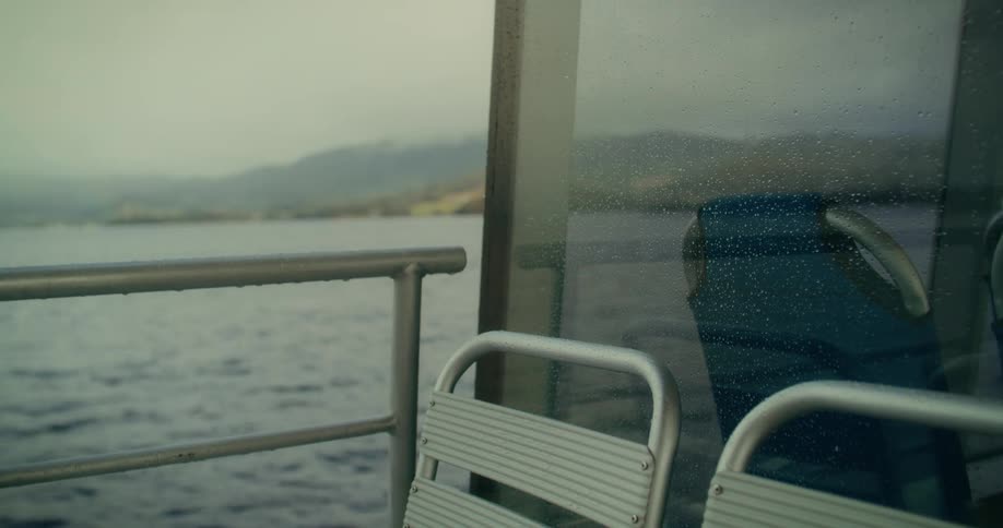 A metal bench on a ferry or boat facing a misty or rainy lake with distant hills