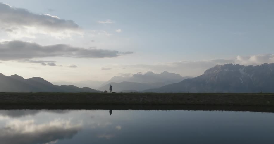 A person and a dog walk on a path by a calm body of water with majestic mountains in the background
