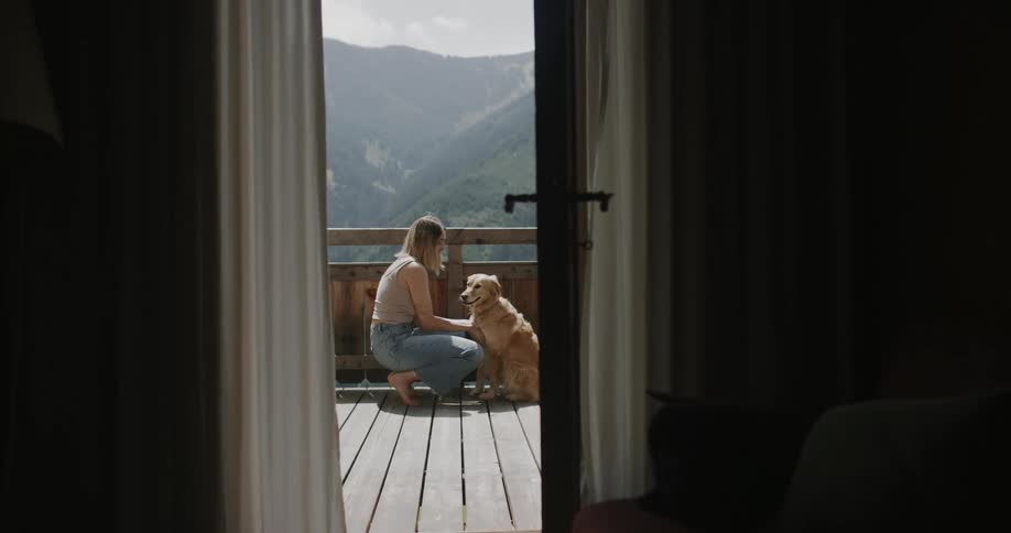 A woman kneels and plays with her golden retriever on a wooden deck with mountains visible in the background through an open door