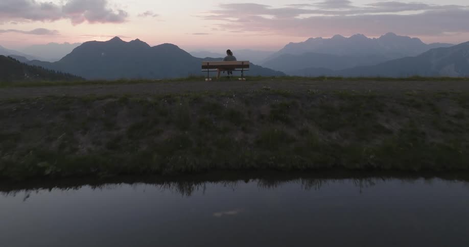 A person sits on a bench overlooking a serene mountain landscape at dusk with a calm body of water in the foreground