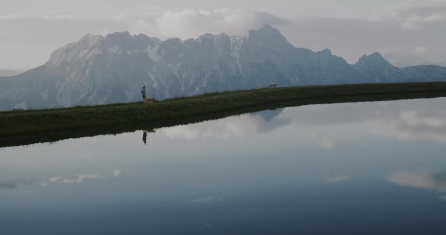 Woman walks with her dog along a narrow strip of grass that stretches over a calm lake reflecting the majestic mountains in the background
