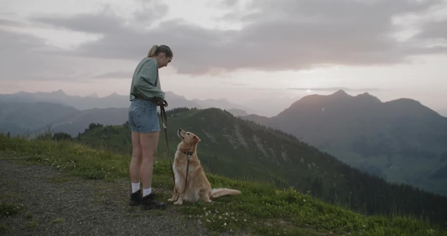 Woman walking her golden retriever on a leash and him sitting in front of her while they are in a space surrounded by mountains