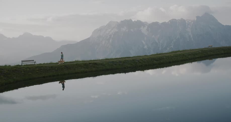 Woman with a dog walking along the edge of a calm lake with the reflection of mountains and sky in the background