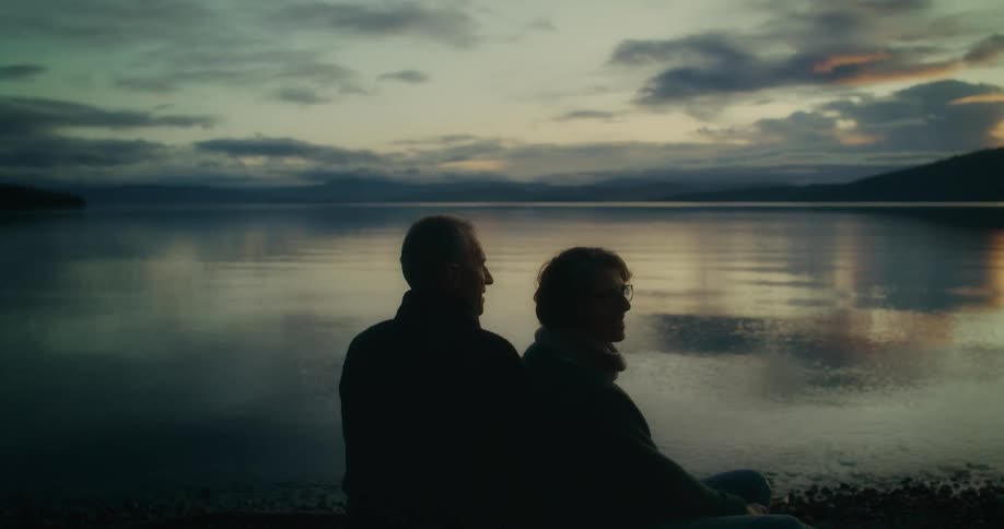 Two people sit by a tranquil lake silhouetted against a serene sunset sky