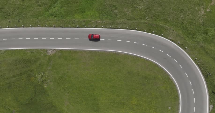 A red car navigates a sharp curve on a winding road surrounded by green grass viewed from above