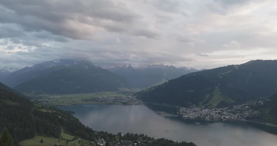 Aerial view of a lake shore town near cloud covered mountains and cows grazing on a hill