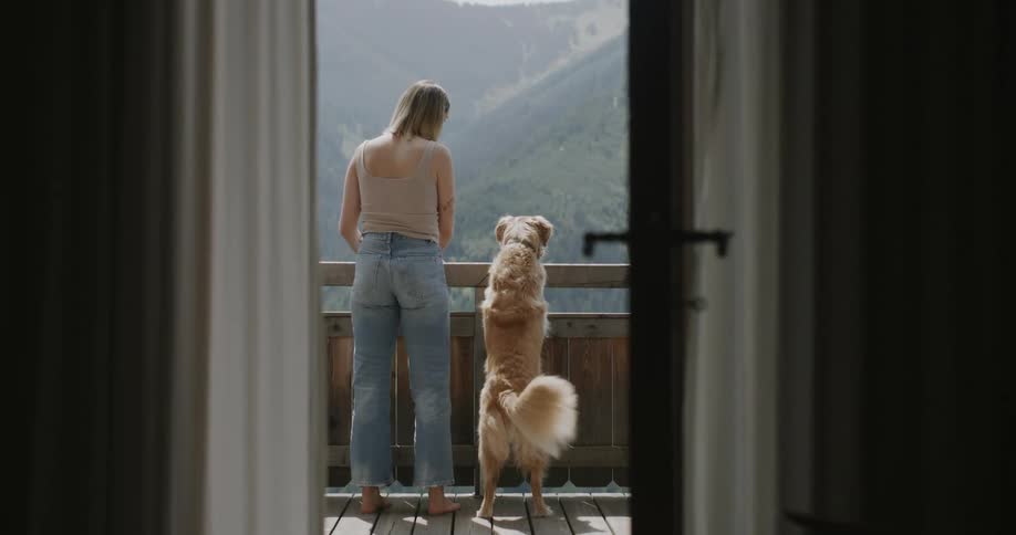A woman and a dog stand on a balcony looking at a mountain range
