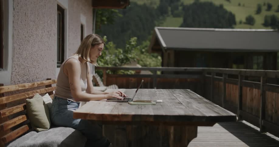 Blonde woman sitting at a desk typing on a laptop while outdoors with mountains in the background
