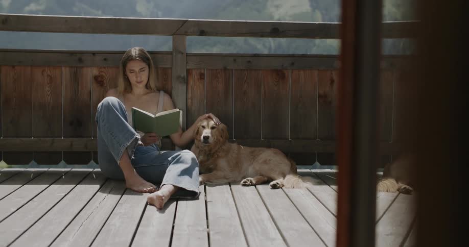 A woman sits on a wooden deck reading a book with her dog lying beside her enjoying a peaceful moment outdoors