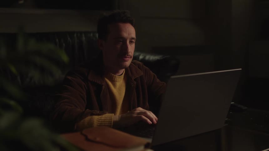 Man is sitting on a sofa typing on a laptop in a lowlight environment