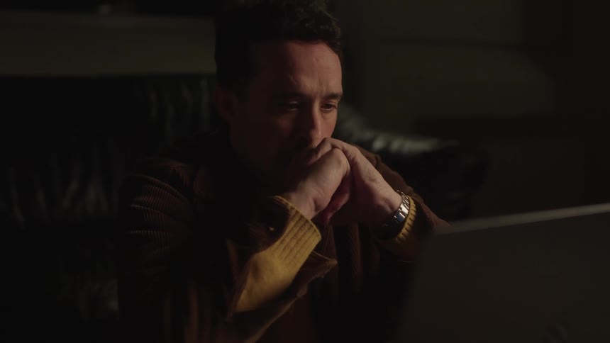 Man is sitting on a sofa crossing his hands over his face in front of a laptop in a lowlight environment