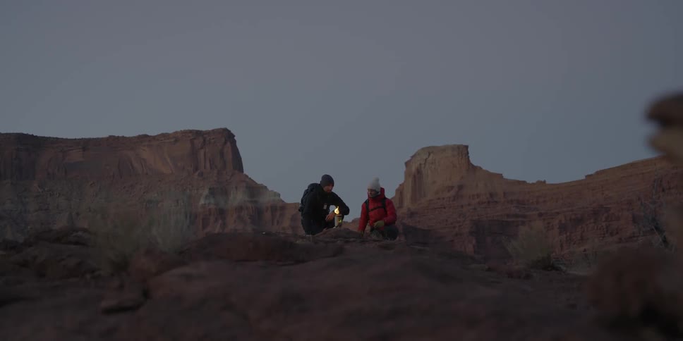 Two people sit near a small fire on a mountain