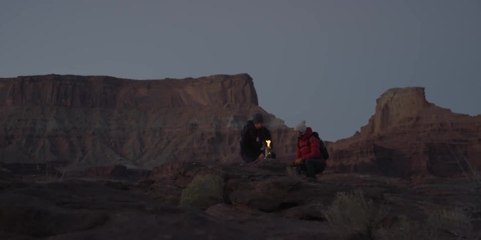 A man and woman sit at a small fire in the mountains