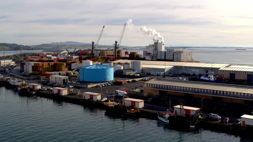 Several boats are docked a shipping yard with a pair of tall cranes