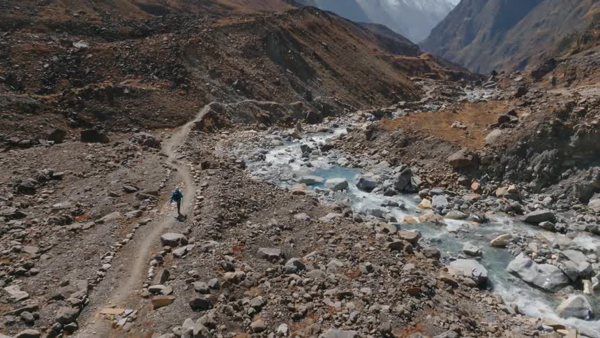 A lone hiker walking along a rocky path beside a mountain stream under clear blue skies