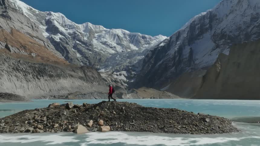 A person is walking on a small rocky island in the middle of a glacial lake surrounded by towering snow covered mountains