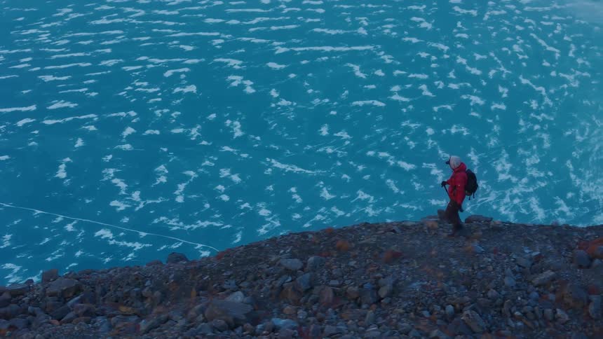 A man wearing a red coat and carrying a backpack walks along a path on the top of a coastal mountain