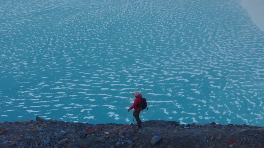 A person in a red jacket walking along a rocky path with a vast body of frozen water filled with floating ice pieces in the background