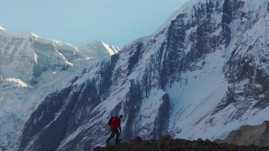 A person in warm clothes is walking on a mountain of rocks and behind there is a mountain covered in snow
