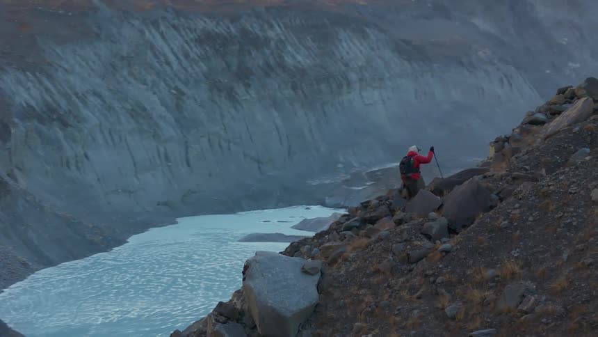 Person climbing a rocky slope with a backpack with a glacial lake and snowcapped mountains in the background