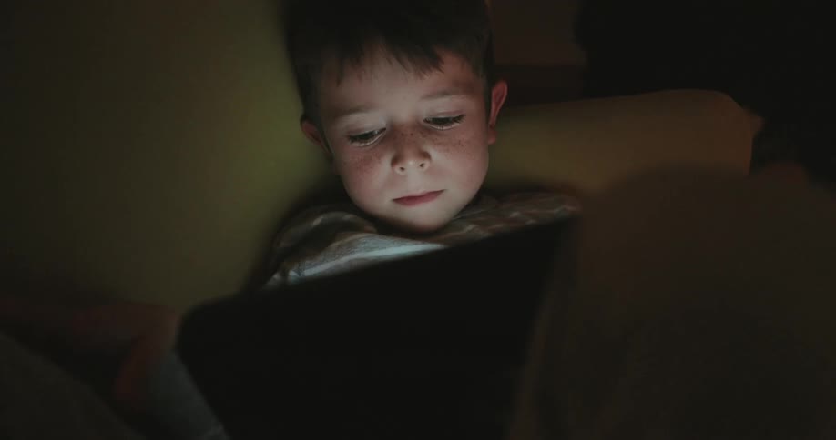 Light from a tablet illuminates the boy watching it in a dark room at night