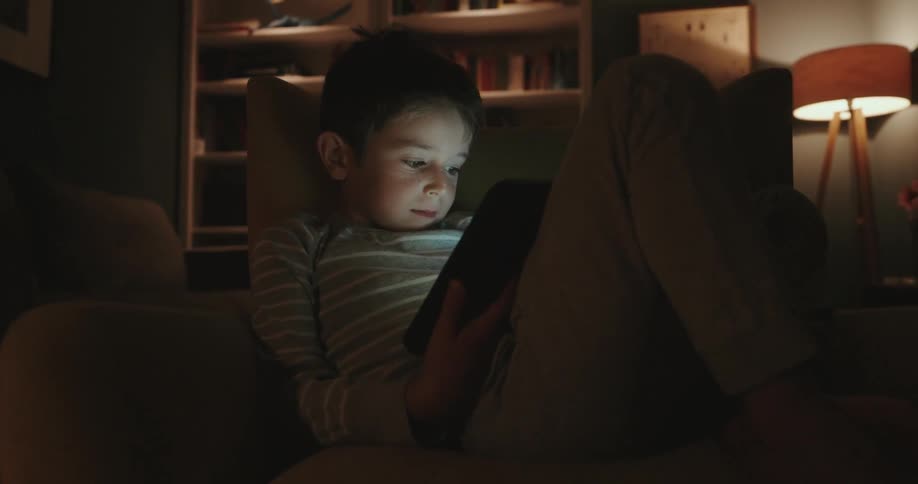 A young boy sits on a couch and reads something on a tablet