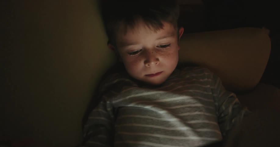 A child sitting in a dimly lit room focused intently on a tablet that illuminates his face