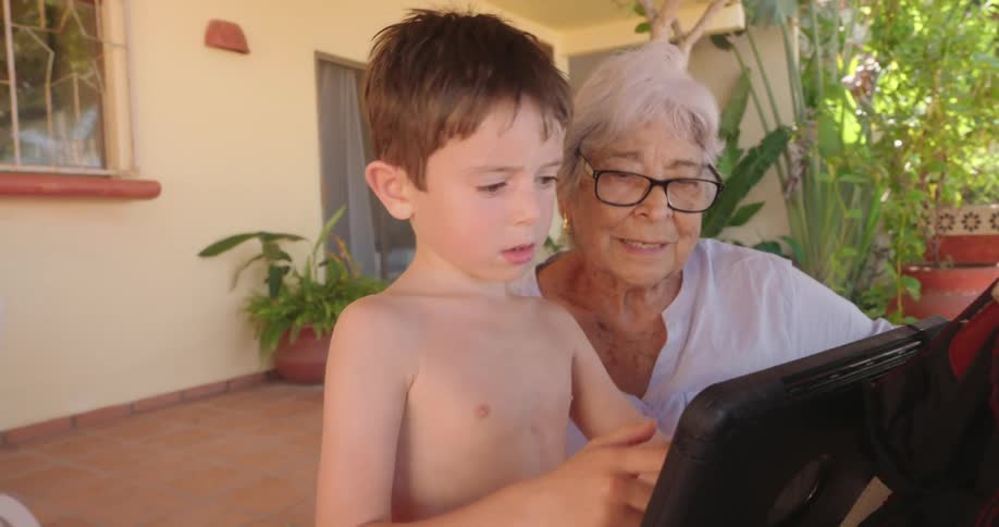 A young boy uses a tablet with his grandmother on the front porch of a house
