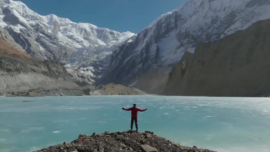A hiker stands on a rocky shore near a frozen lake and huge mountains with arms spread wide