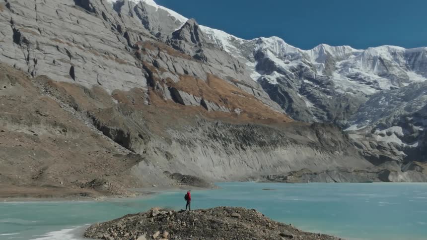 Person in the center of a rock surrounded by a blue lake and mountains with snow around