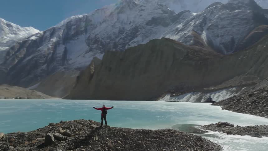 Backpacker stands on a mound near a frozen lake with their arms spread wide as they face the snowy mountains