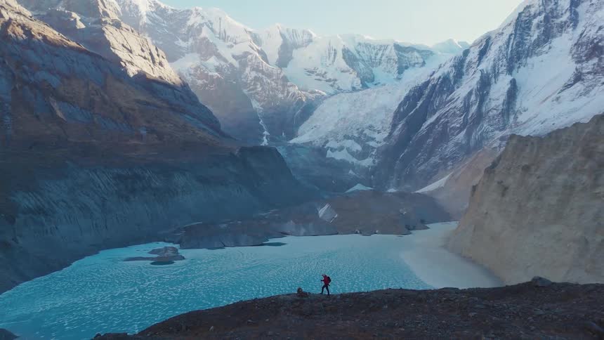 A man hikes across a steep rocky slope in front of a glacial lake framed by towering snow covered mountains