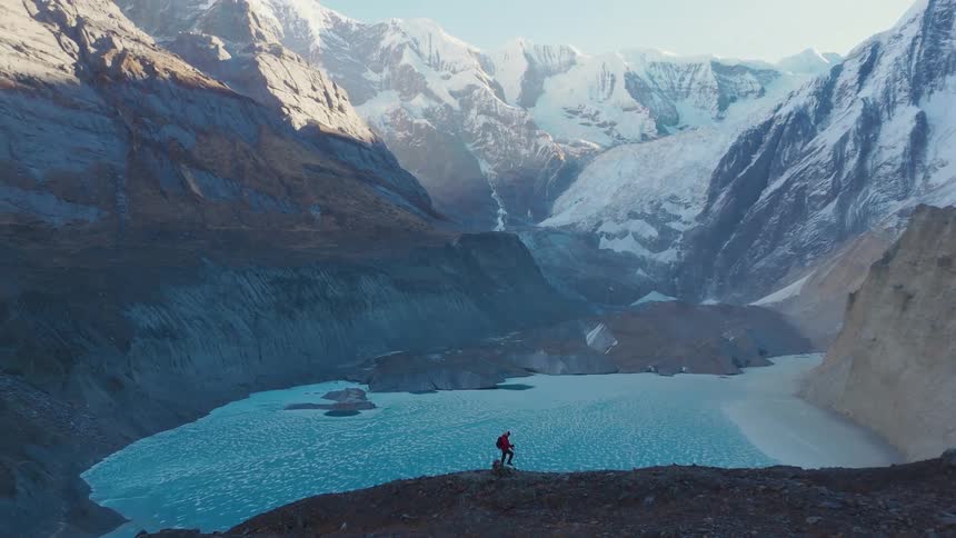 A man hikes along a steep rocky slope in front of a mountainous landscape with a serene glacial lake