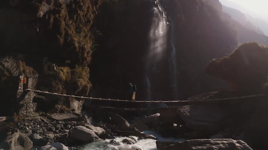 Person crossing a wooden bridge above a river with mountains and a waterfall around