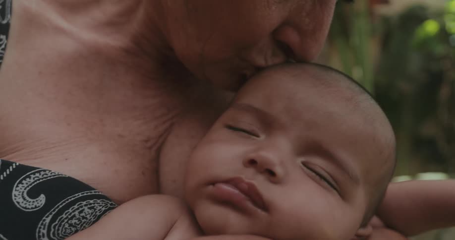 An older woman holds a baby outside of a house