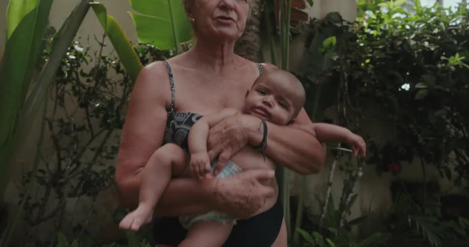 An older woman in a swimsuit rocks a baby in her arms while being outside a home
