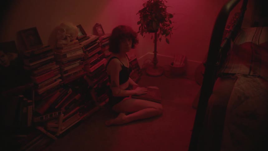 A woman in her bra and underwear sits on a bedroom floor