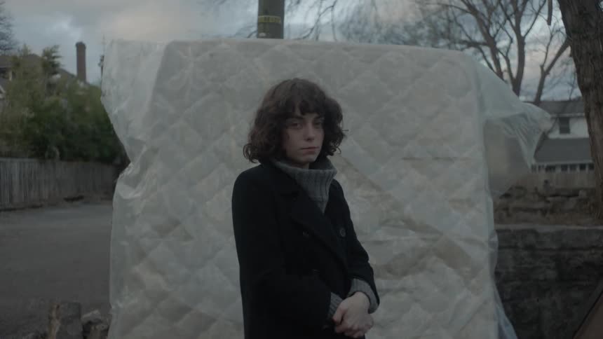 A woman wearing a coat is standing near a mattress outdoors