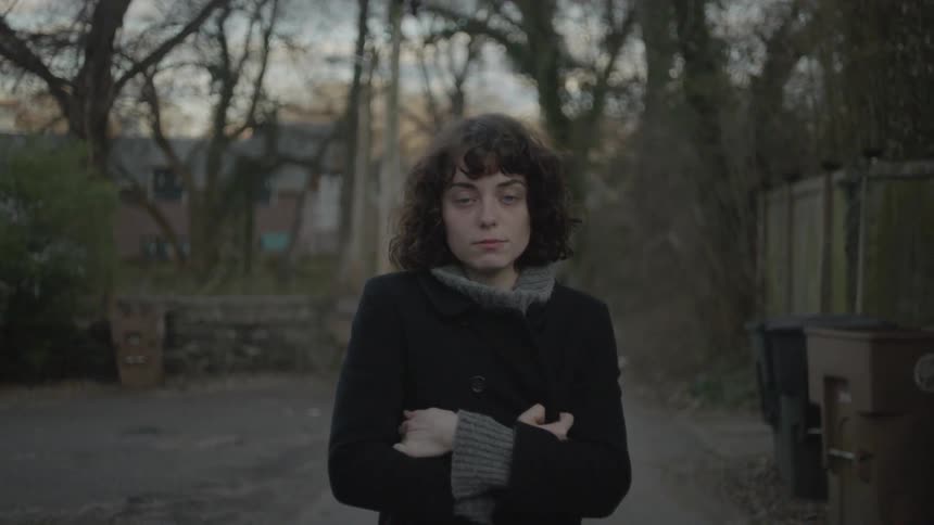 A woman in a coat stands outside with trees in the background